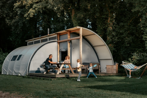 Gezin voor Arcade-lodge: ouders aan picknicktafel, twee spelende kinderen, één kind in hangmat rechts.