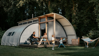 Familie nyder tid foran Arcade-lodge. To børn leger, voksne sidder ved bord, hængekøje til højre.