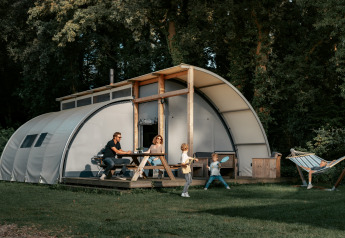 Gezin voor Arcade-lodge: ouders aan picknicktafel, twee spelende kinderen, één kind in hangmat rechts.