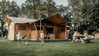 Gezin geniet van buitenleven bij een grote Safari Lodge tent, kinderen spelen op het gras ervoor.