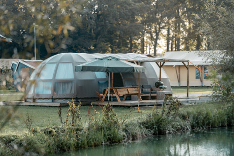 Panorama lodge met moderne koepeltent, terras met parasol en uitzicht op het meer en het bos.