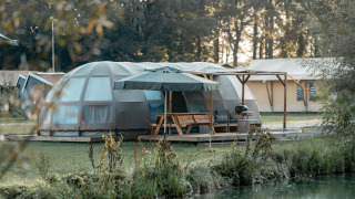 Panorama lodge met moderne koepeltent, terras met parasol en uitzicht op het meer en het bos.