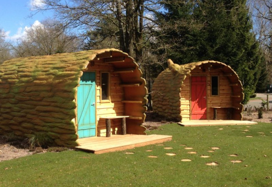 Dos acogedoras cabañas con forma de tronco de árbol y puertas de colores rodeadas de naturaleza y césped.