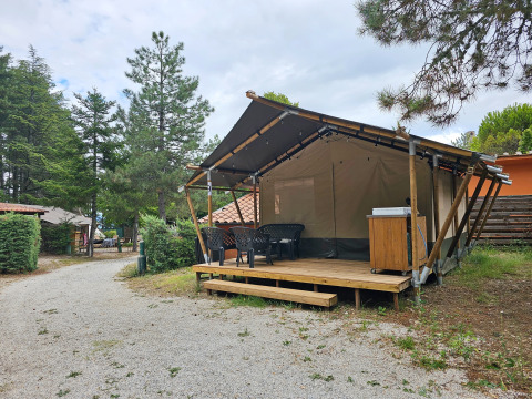 Safari tent on a wooden deck with plastic chairs and table, surrounded by trees and a gravel path.
