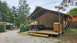 Safaritent op een houten vlonder met plastic stoelen en tafel, omgeven door bomen en een grindpad.
