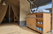 Vista interior de una tienda safari con suelo de madera, cocina y comedor en Camping Luna del Monte, Italia.