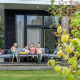 Børn sidder på en terrasse foran River Cabin på Holiday Park Mölke, omgivet af grønt og leger.