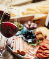 Dos copas de vino tinto y una tabla de embutidos con pan, carnes y aceitunas en un parque de glamping.