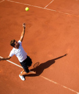 Tennis player serving the ball on a clay court at a holiday park with glamping, seen from above.