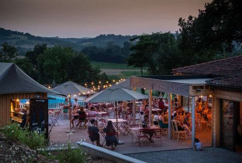 Terraza con mesas, sombrillas y gente cenando al atardecer en Huttopia Pays de Cordes sur Ciel, Occitanie, Francia.