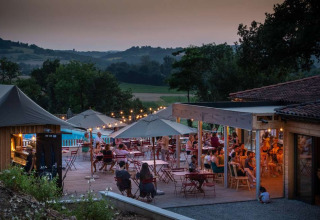 Udendørs restaurantterrasse ved solnedgang med gæster, parasoller og lys på Huttopia Pays de Cordes sur Ciel i Occitanie.