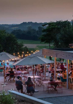 Terraza con mesas, sombrillas y gente cenando al atardecer en Huttopia Pays de Cordes sur Ciel, Occitanie, Francia.