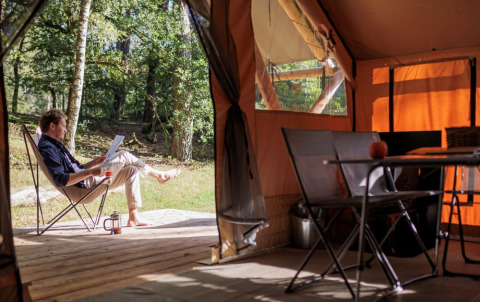 Hombre relajándose con libro y café frente a una tienda en Camping Huttopia De Veluwe, Gelderland, Países Bajos.