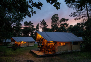 Beleuchtete Zelte im Camping Huttopia De Veluwe, umgeben von Bäumen bei Sonnenuntergang in Gelderland.