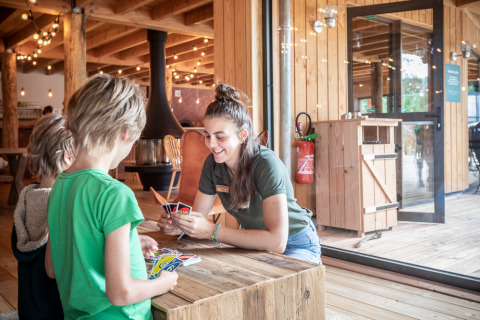 Niños juegan a las cartas con una adulta en el interior de una cabaña de madera en Camping Huttopia De Veluwe.