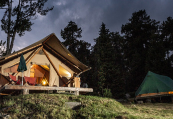 La tente safari Canadienne Cosy State illuminée le soir, entourée d’arbres et d’une autre tente verte en forêt.