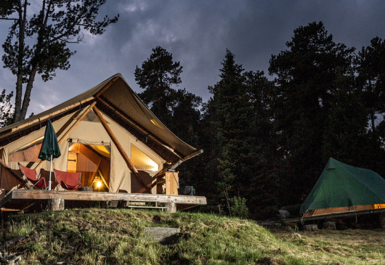 Das Safari-Zelt Canadienne Cosy State abends beleuchtet, umgeben von Bäumen und einem grünen Zelt im Wald.