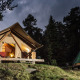 Tenda safari Canadienne Cosy State illuminata di sera, circondata da alberi e una seconda tenda verde nel bosco.