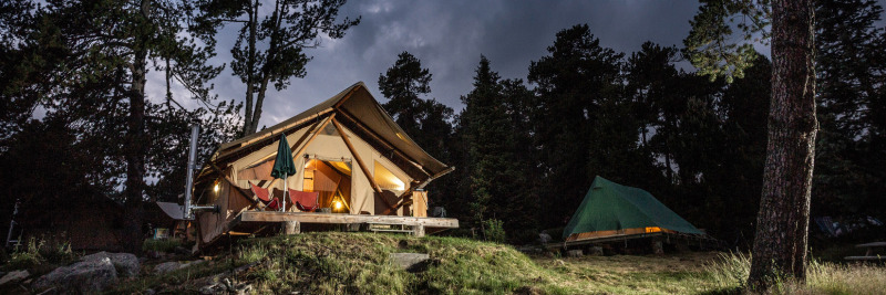 Das Safari-Zelt Canadienne Cosy State abends beleuchtet, umgeben von Bäumen und einem grünen Zelt im Wald.