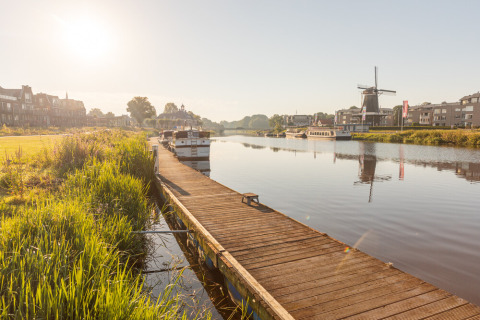 Zonnige rivier met boten en houten steiger, windmolen op de achtergrond bij Huttopia De Roos in Overijssel.