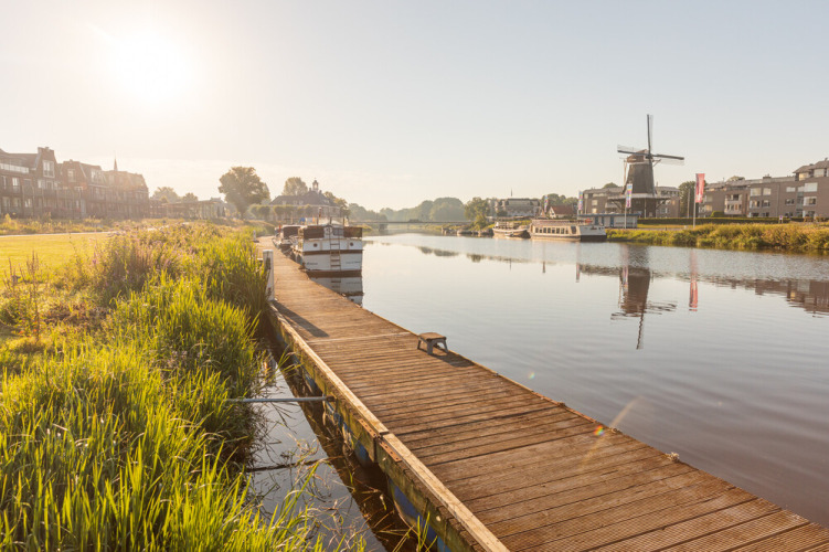 Sunny riverside with boats, a wooden dock, and a windmill at Huttopia De Roos holiday park in Overijssel, Netherlands.