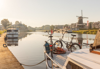 To cykler på en båd ved en rolig flod med en vindmølle og både i det fjerne på Huttopia De Roos, Holland.