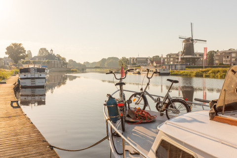 Deux vélos sur un bateau amarré à une rivière calme avec moulin à vent et bateaux à Huttopia De Roos, Pays-Bas.