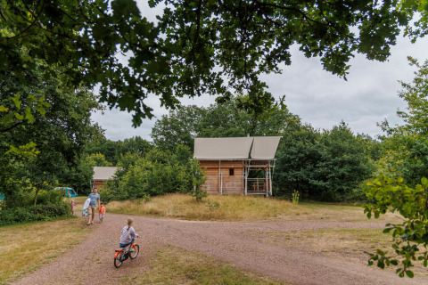 Gezin fietst bij houten huisjes in het bosrijke vakantiepark Huttopia De Roos in Overijssel, Nederland.