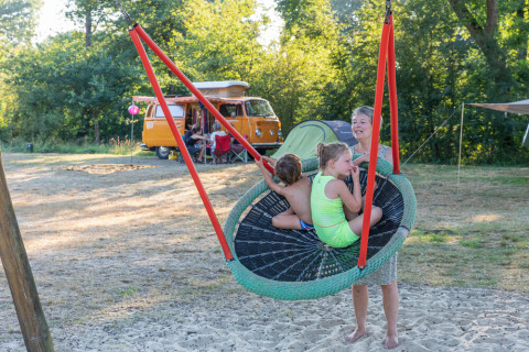 Familia disfruta de un columpio grande en un camping con furgoneta VW amarilla y tienda de fondo en Overijssel.