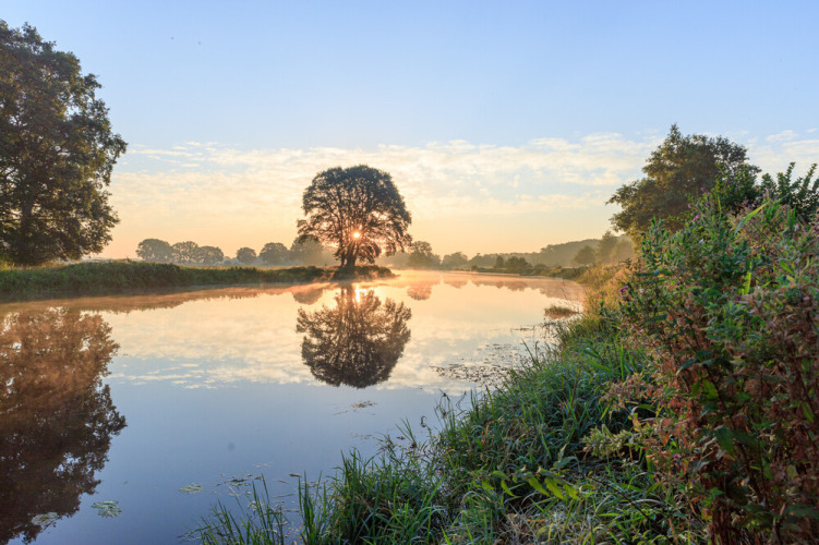 Zonsopgang aan een rustige rivier met boomreflecties bij Huttopia De Roos, Overijssel, Nederland.