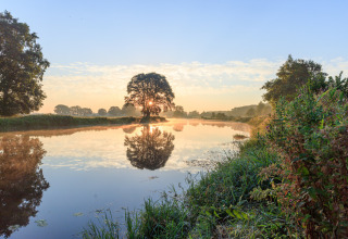 Sonnenaufgang über einem ruhigen Fluss mit Baumspiegelung bei Huttopia De Roos, Overijssel, Niederlande.