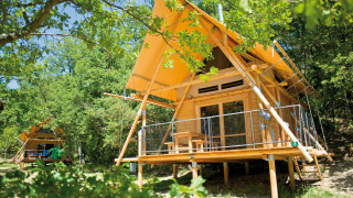 Tienda safari Cahutte con techo amarillo y terraza de madera, rodeada de bosque verde y frondoso.