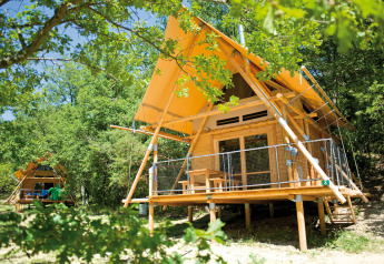 Tienda safari Cahutte con techo amarillo y terraza de madera, rodeada de bosque verde y frondoso.