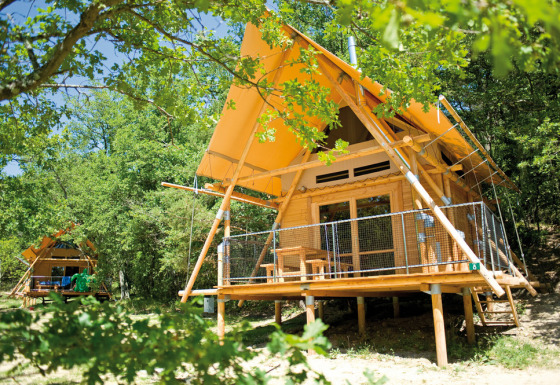 Tienda safari Cahutte con techo amarillo y terraza de madera, rodeada de bosque verde y frondoso.