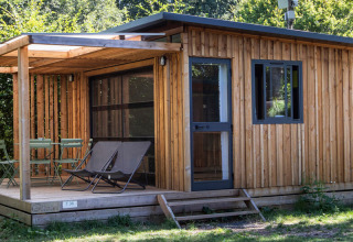 Træbeklædt chalet med overdækket terrasse og havemøbler, omgivet af frodige grønne omgivelser ved Huttopia De Roos.