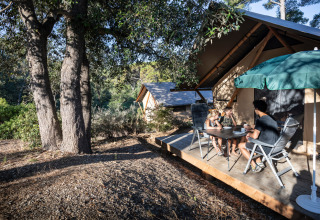 Familia disfruta afuera de una tienda safari Trappeur en Huttopia Lac de l'Uby - Gers, Francia, entre árboles.
