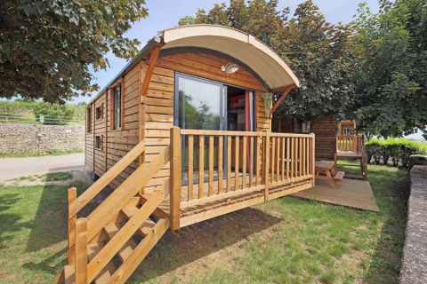 Houten bungalow genaamd Gypsy caravan met veranda en treden, tussen bomen en gras in het zonlicht.