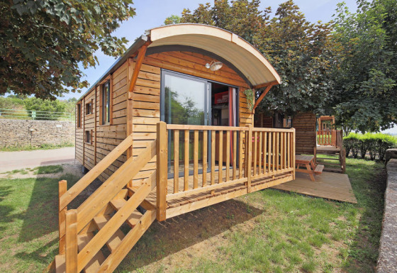 Houten hut genaamd Gypsy caravan met veranda en trap, omgeven door gras en bomen in de zon.