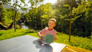 Niño jugando al ping pong al aire libre bajo el sol en un parque vacacional que ofrece glamping.