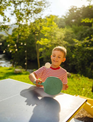 Dreng, der spiller bordtennis i det fri med grønne omgivelser og solskin på en feriepark med glamping.