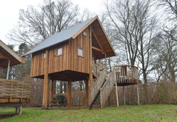 Casa sull’albero chiamata Ooievaarsnest con scala in legno e terrazza, circondata da alberi e prato.