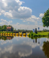 Vista panorámica de un lago con reflejos, árboles verdes y una zona de glamping bajo el cielo azul.