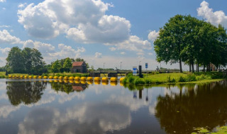 Panoramisch uitzicht op een meer met weerspiegeling, groene bomen en glamping onder een blauwe lucht.