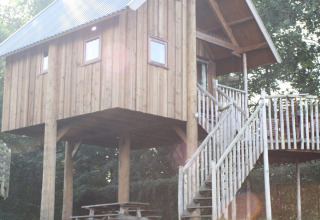 A wooden tree house on stilts with terrace and stairs, bathed in sunlight, at Camping de Koeksebelt, Netherlands.