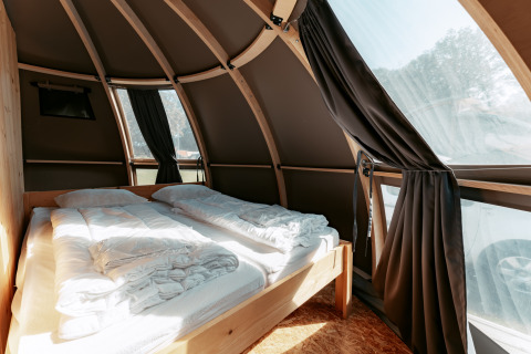 Vista interior de una moderna cabaña curva con cama y grandes ventanales en Camping de Italiaanse Meren.