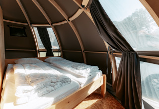 Vista interior de una moderna cabaña curva con cama y grandes ventanales en Camping de Italiaanse Meren.