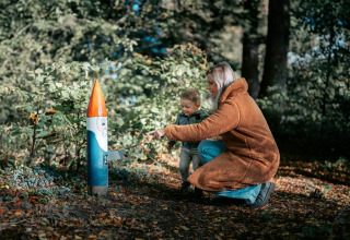 En kvinde og et lille barn udforsker skoven nær en farverig træfigur ved Camping de Italiaanse Meren.