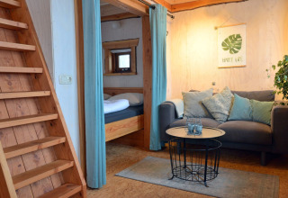 Gezellige zithoek met sofa en houten trap in de boomhut Ooievaarsnest bij Camping de Koeksebelt, Nederland.