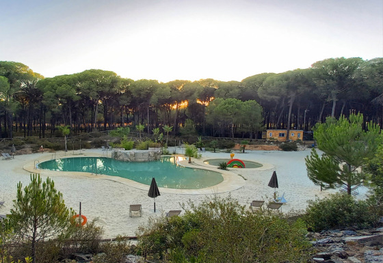 Zicht op een zwembad met zand omgeven door bos bij zonsondergang in Huttopia Parque de Doñana, Andalusië.