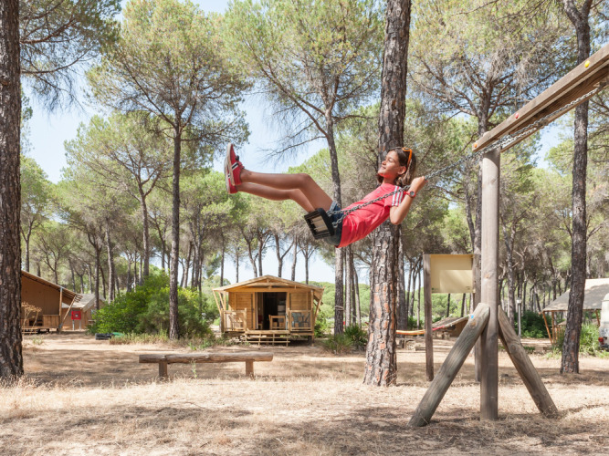 Niño columpiándose en parque de juegos en bosque de pinos, cerca de cabañas en Huttopia Parque de Doñana.